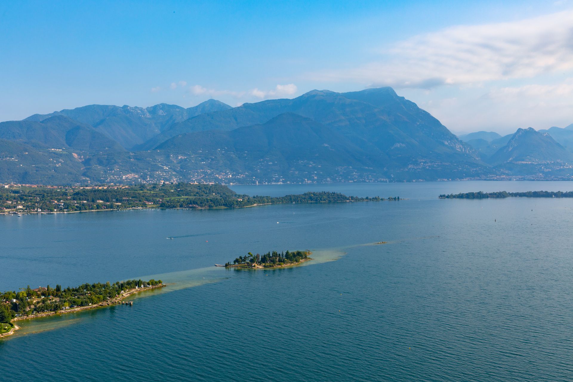 Veduta aerea di un lago blu con una piccola isola e una penisola, incorniciato da una catena montuosa sotto un cielo azzurro e limpido.