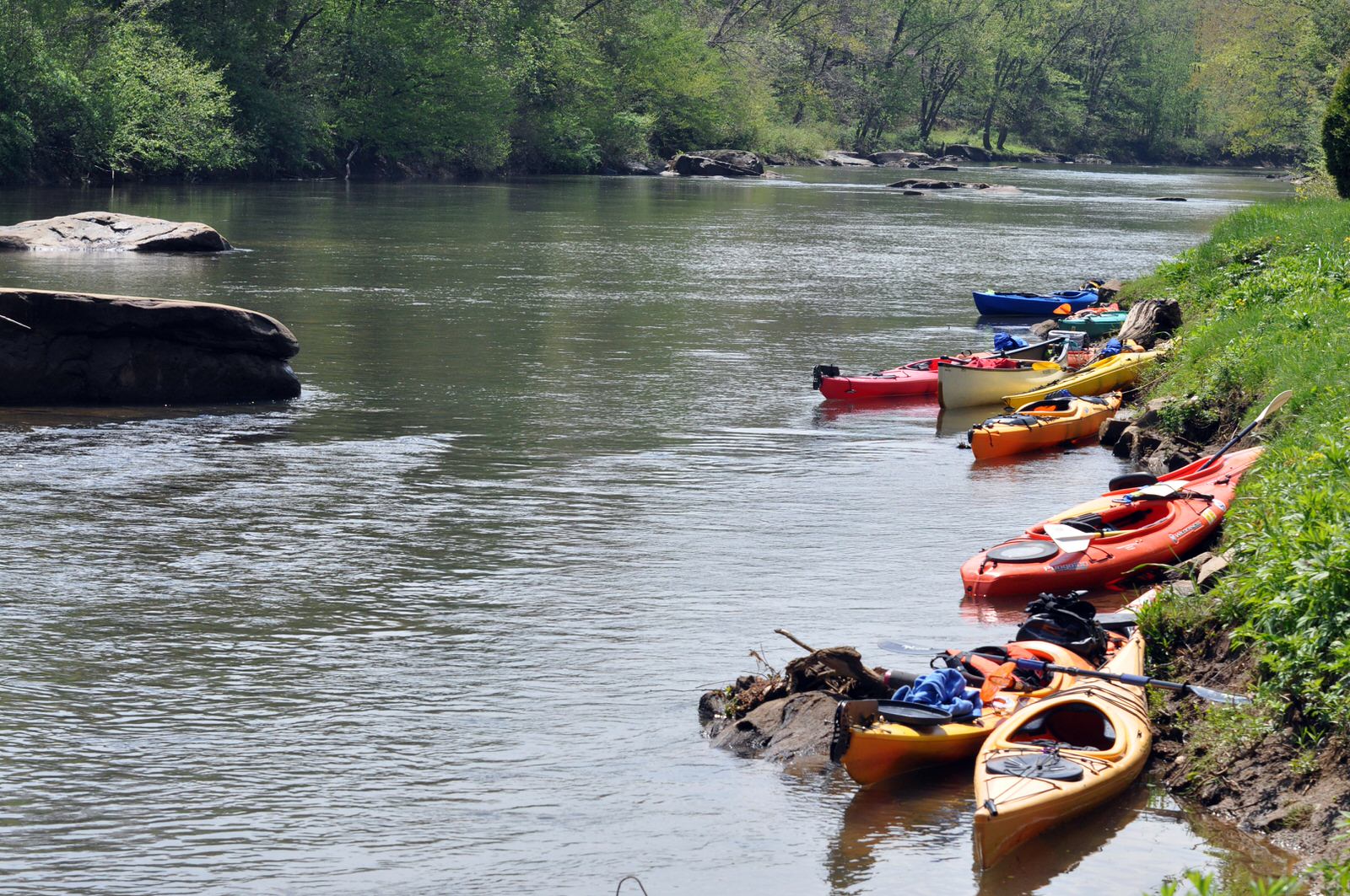 Kayaks line the muddy riverbank; green trees frame the flowing water on a sunny day.