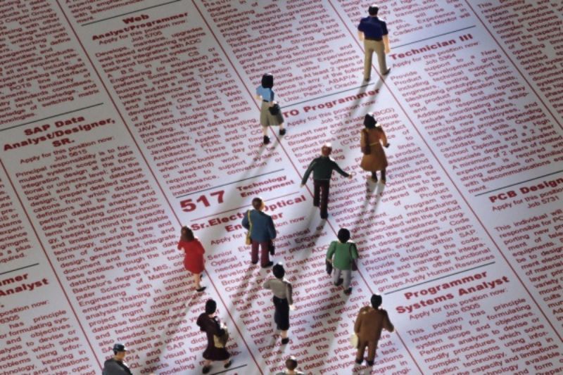 Miniature people standing on a newspaper classifieds section, searching for job openings.
