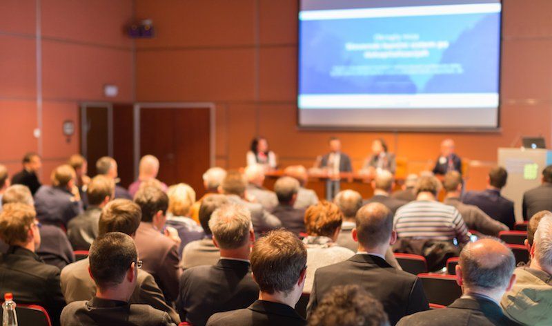 Audience at a conference listening to panelists at a table on a stage with a projection screen.