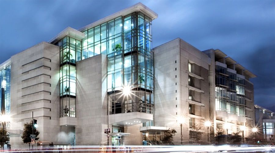Modern, light-colored building with glass tower sections, lit up at dusk; street in the foreground.