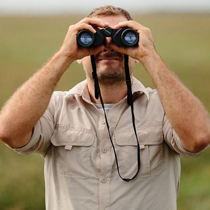 A man in a tan shirt is looking through binoculars
