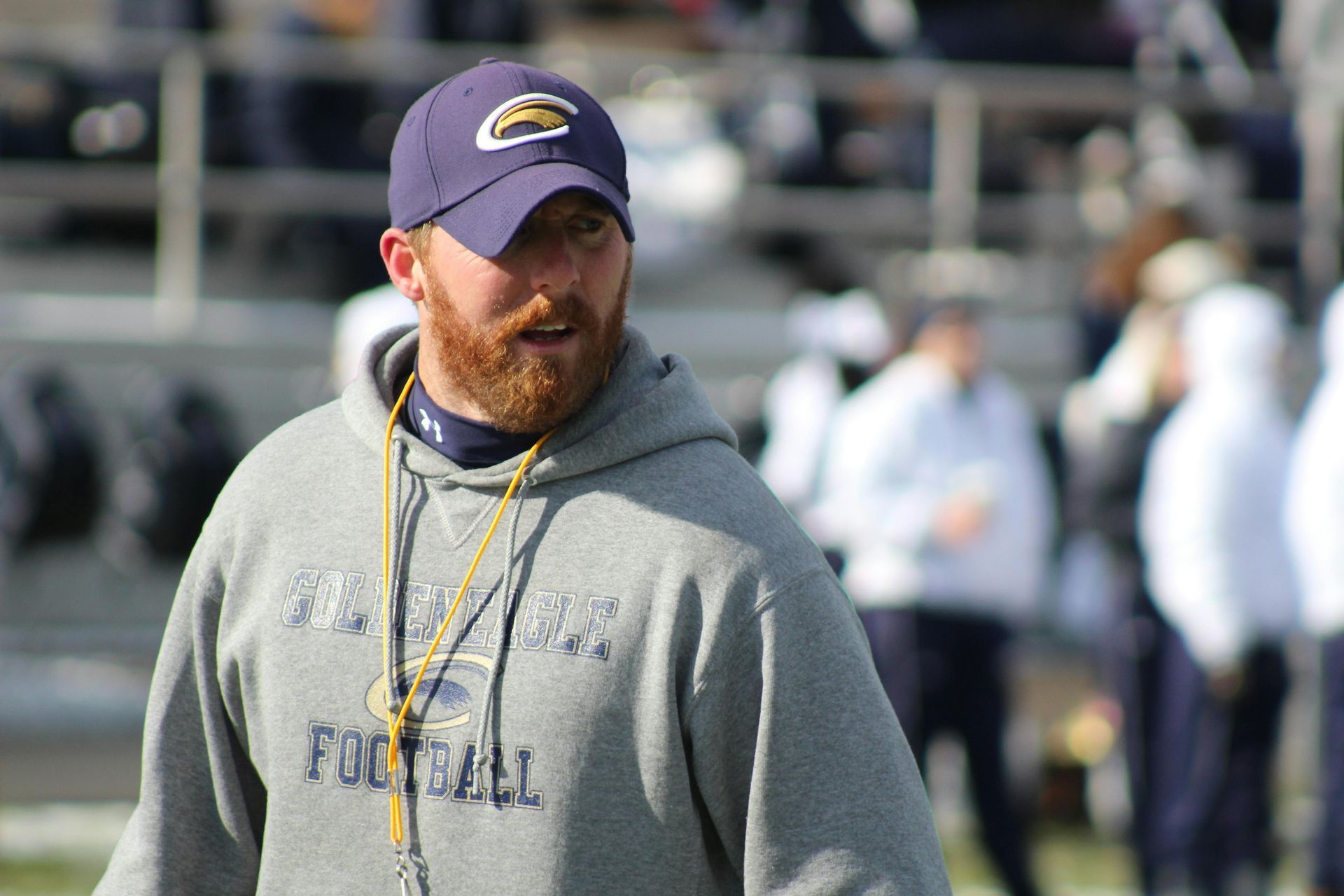 Man in navy cap and grey hoodie coaches football on a field.
