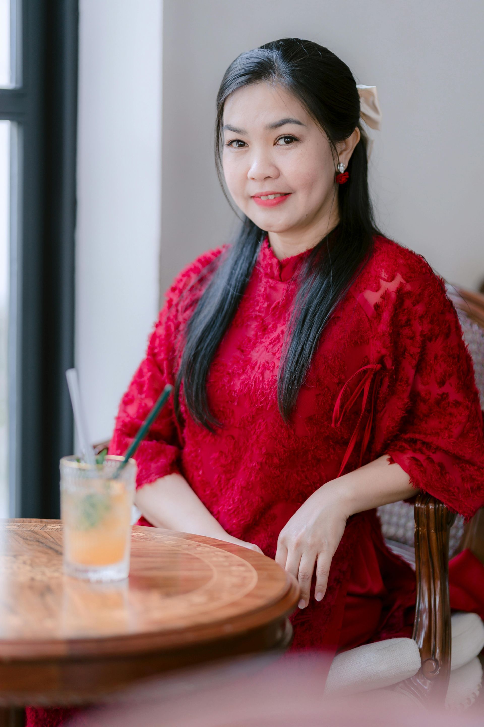 Woman in red dress, seated at a table with a drink, smiles indoors.
