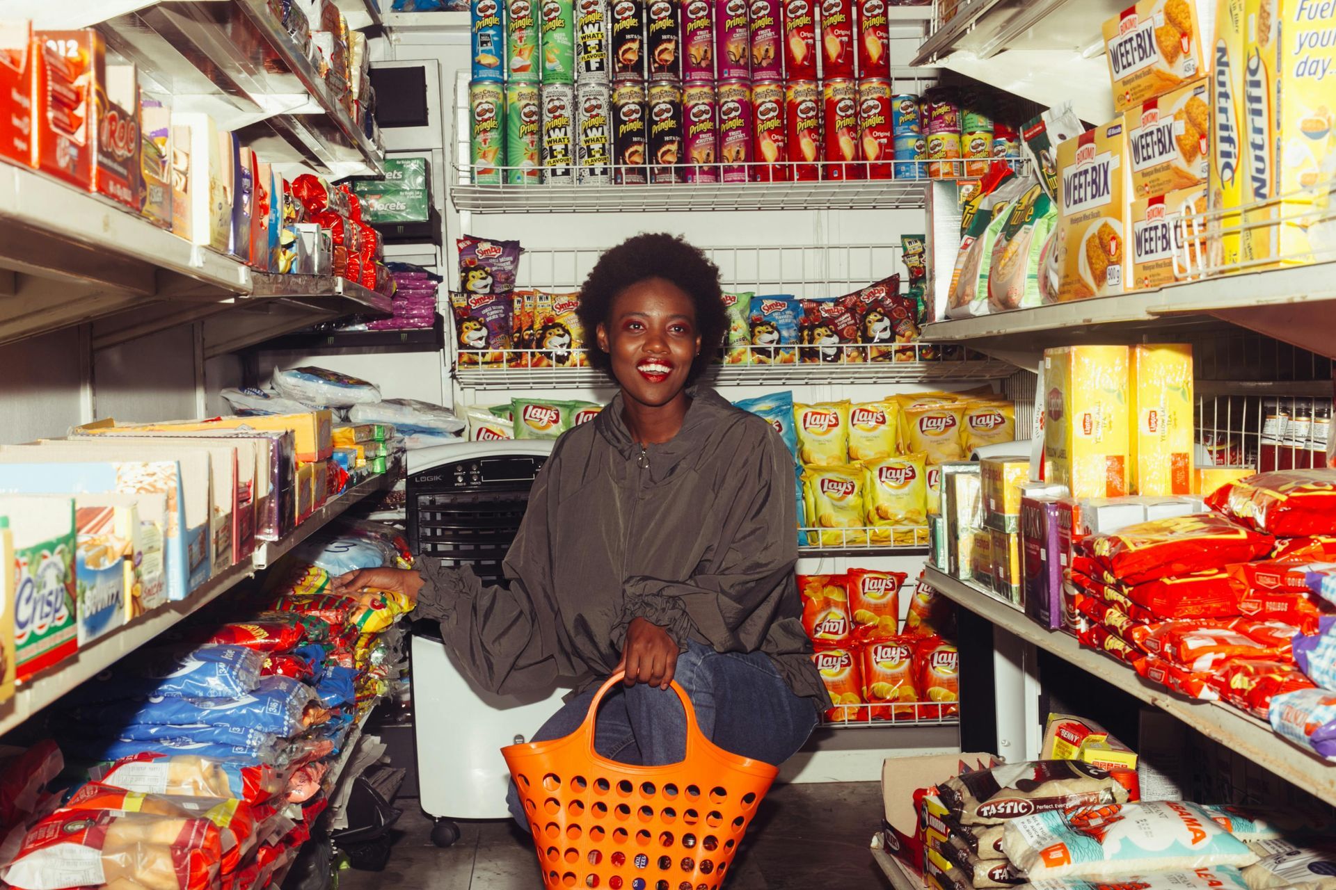 Woman in a small grocery store smiling, holding a basket. Surrounded by snacks on shelves.