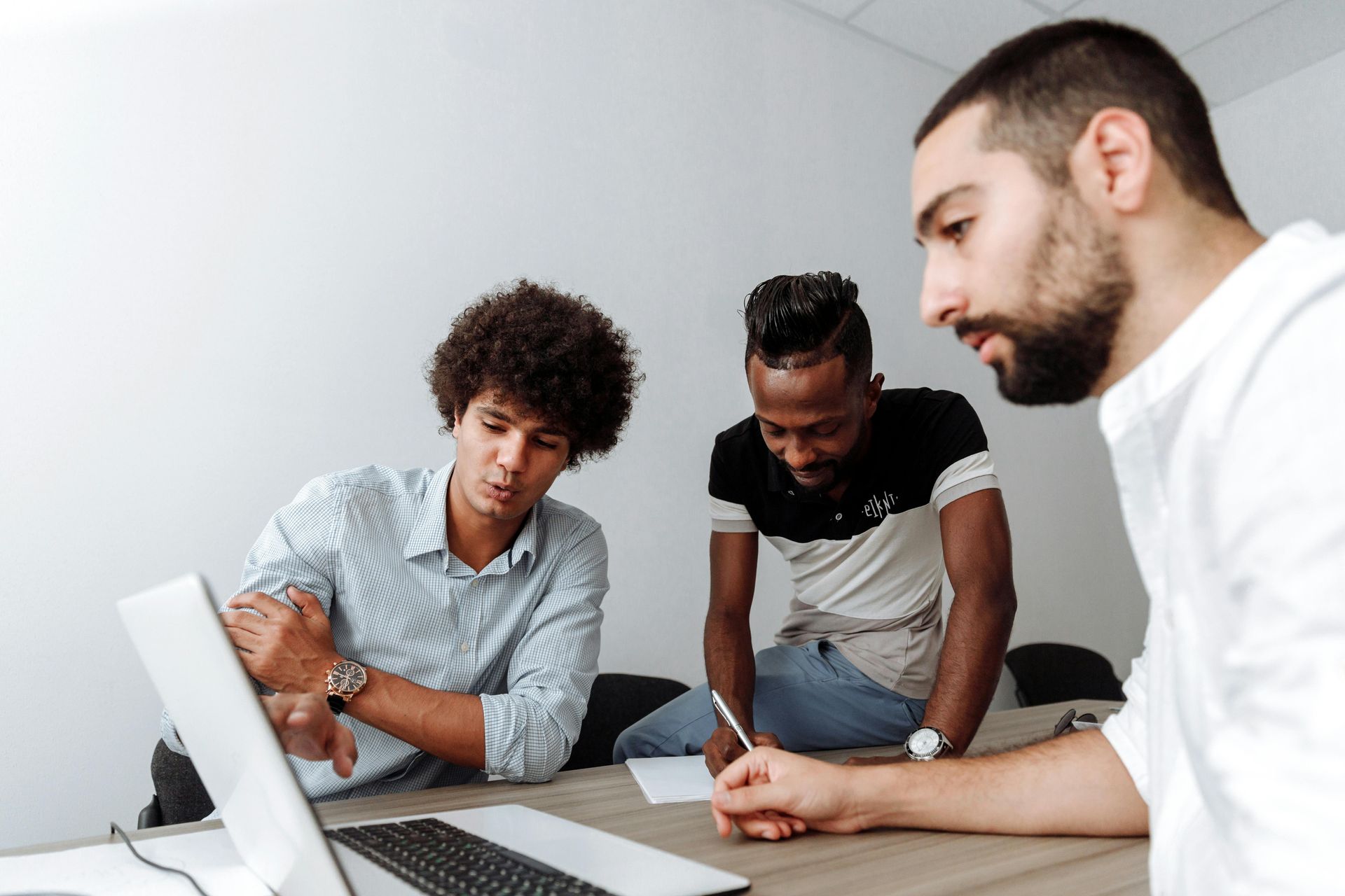 Three men collaborating at a desk, focused on a laptop. One points, another takes notes. Plain white room.