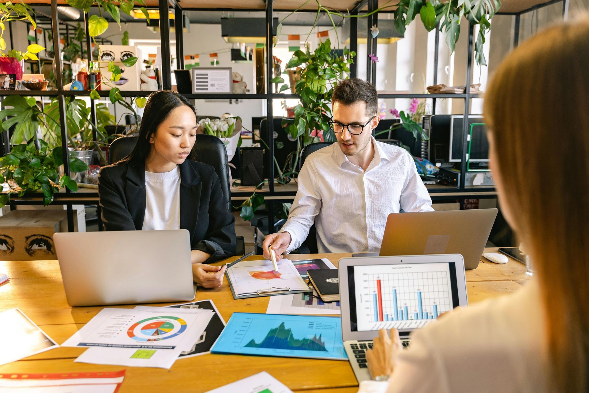 Three people in an office, working on laptops and reviewing charts on a wooden table. Green plants in background.