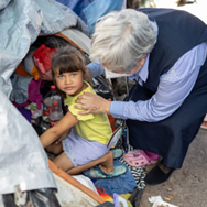 Woman comforts a child amidst cluttered items; both are outdoors. The child looks toward the camera.