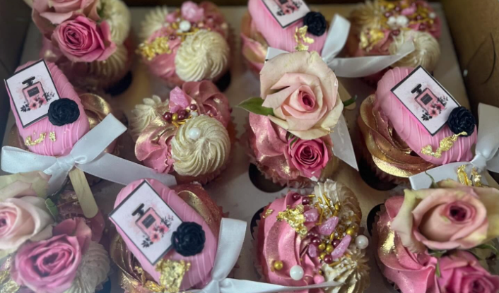 A box of pink and gold cupcakes with flowers on them.