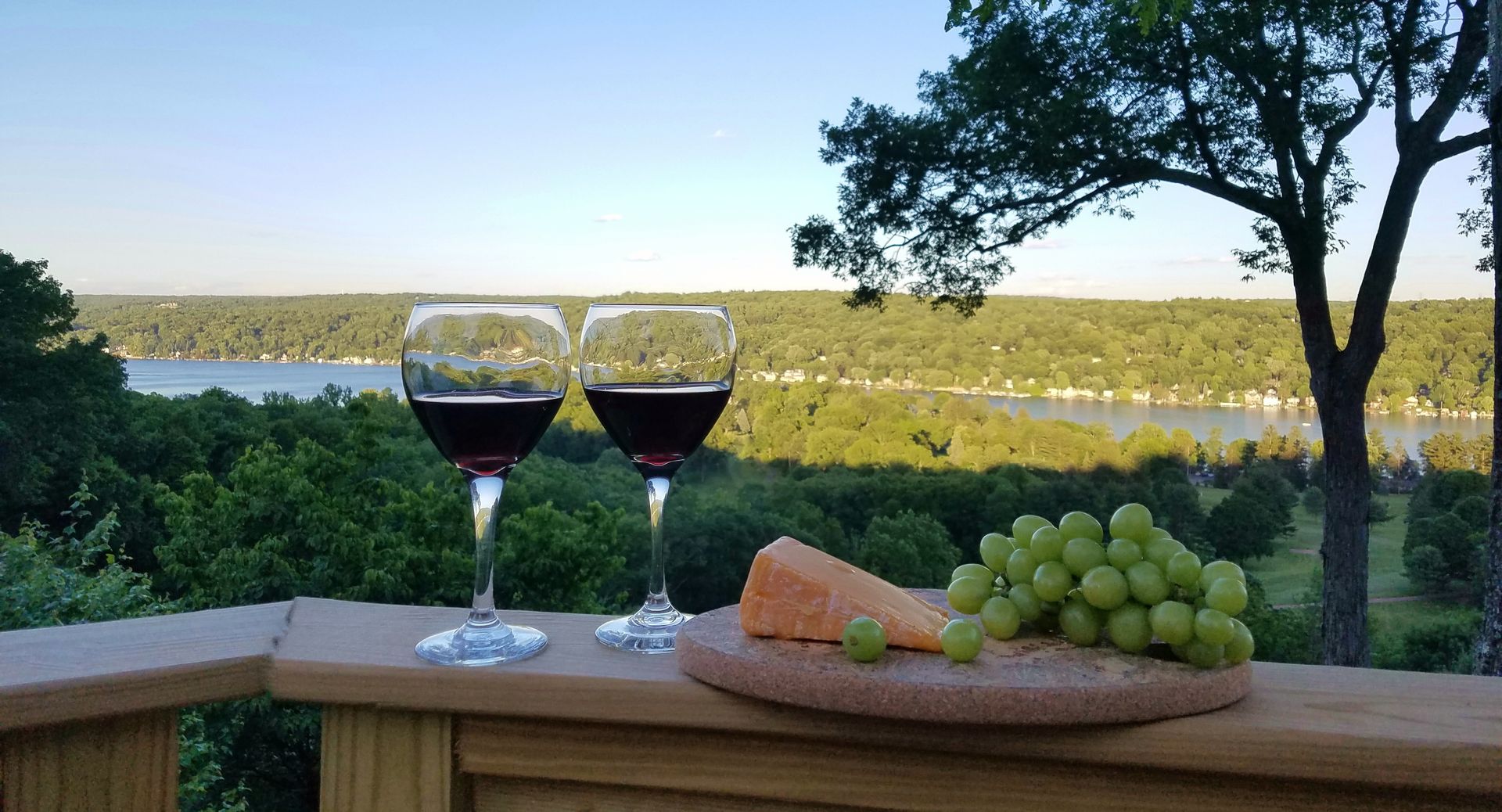 Two glasses of wine and grapes on a wooden deck overlooking a lake.