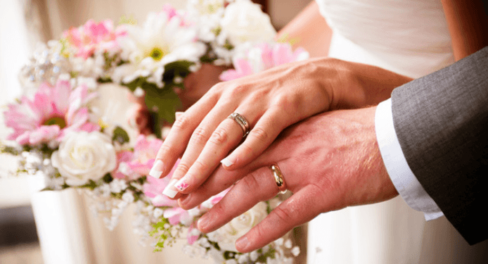 A bride and groom are putting their wedding rings on their hands while holding a bouquet of flowers.