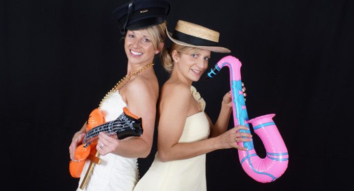 Two women are standing back to back holding ukuleles and a pink saxophone.