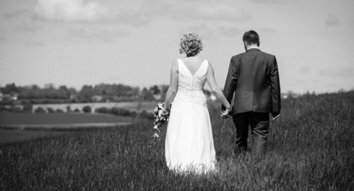 A bride and groom are walking through a field holding hands.