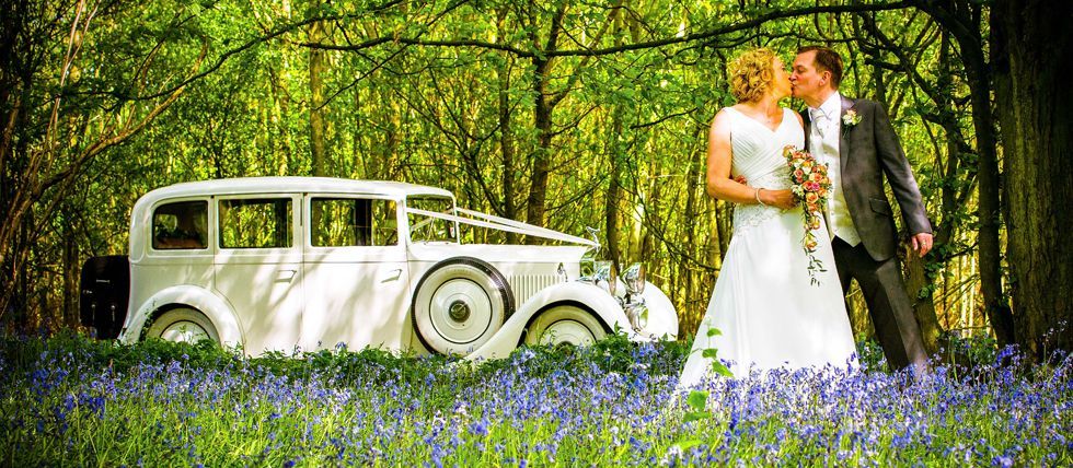 A bride and groom are kissing in front of a white car in the woods.