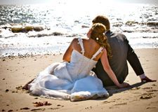 A bride and groom are sitting on the beach looking at the ocean.