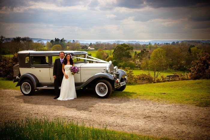 A bride and groom are posing for a picture in front of an old fashioned wedding car.