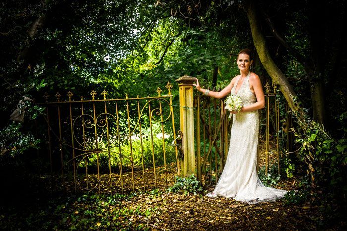 A bride in a wedding dress is standing in front of a gate in the woods.