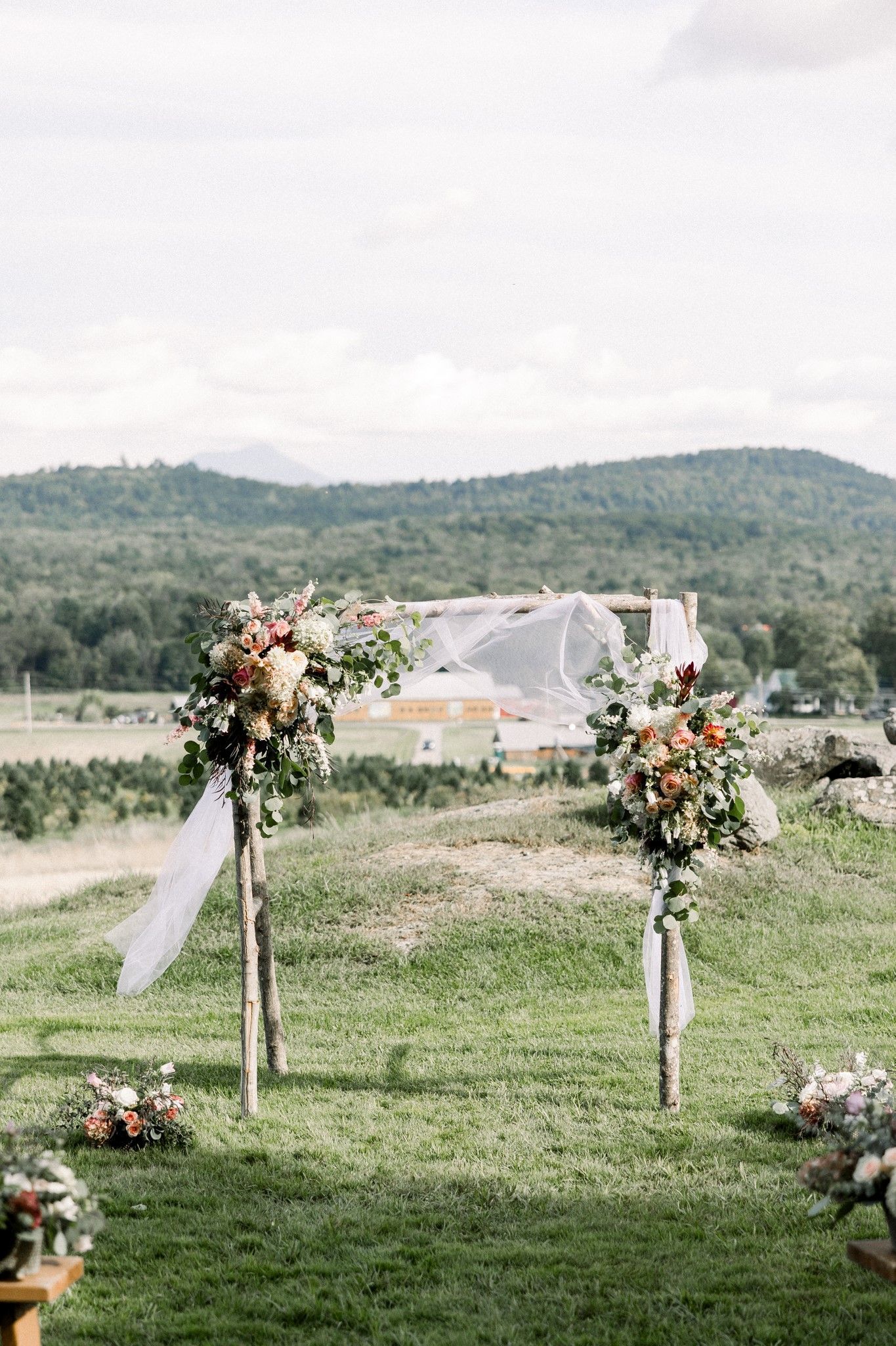 wedding details shot of rustic ceremony arch draped with sheer white fabric and floral arrangements