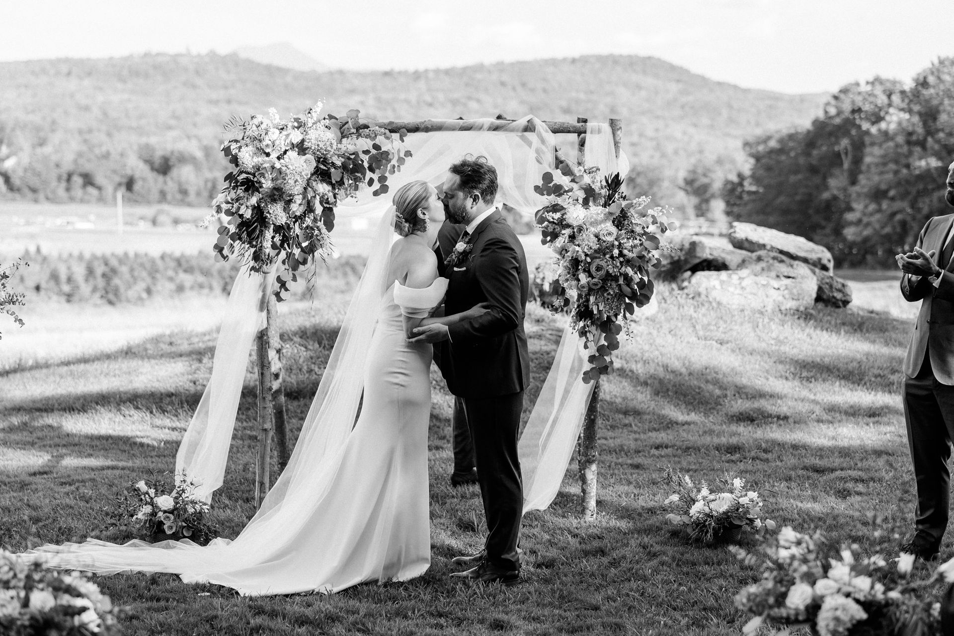 black and white photo of a newlywed couple sharing their first kiss in front of their ceremony arch at isham family farm