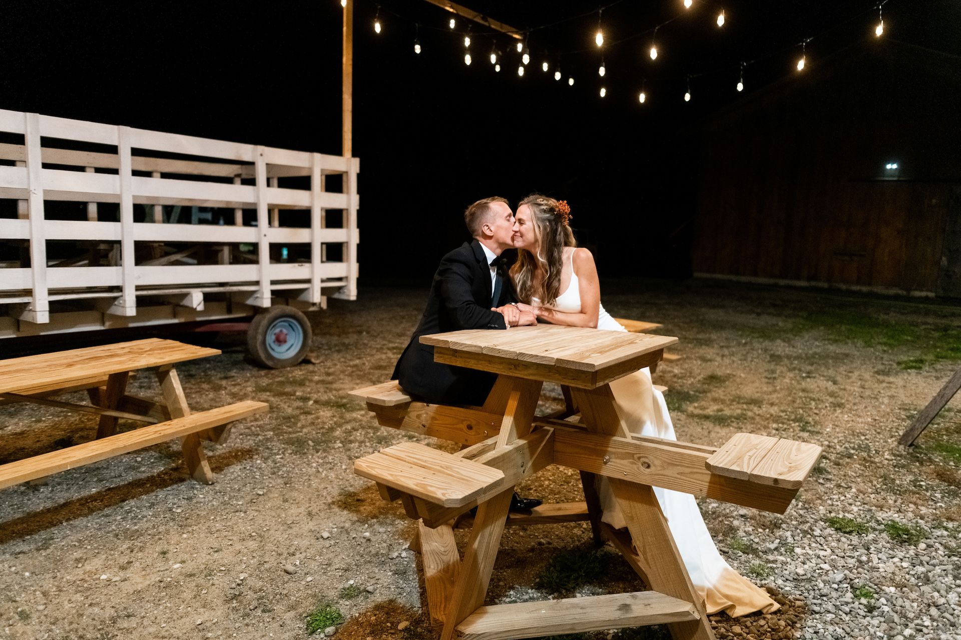 landscape photo of newlywed couple kissing on a wooden bench outside of their reception at isham family farm events barn in vt