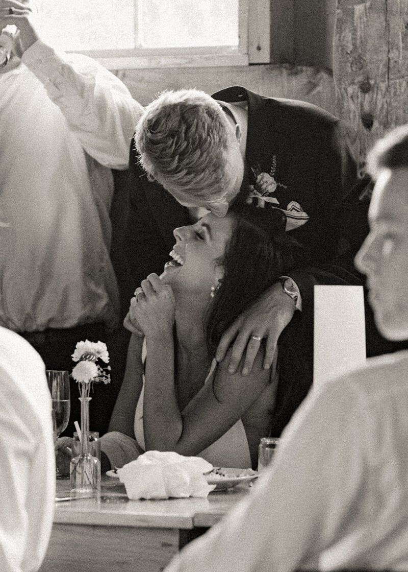 Groom kisses bride’s head as she laughs during reception—a candid black-and-white shot full of warmth and joy.
