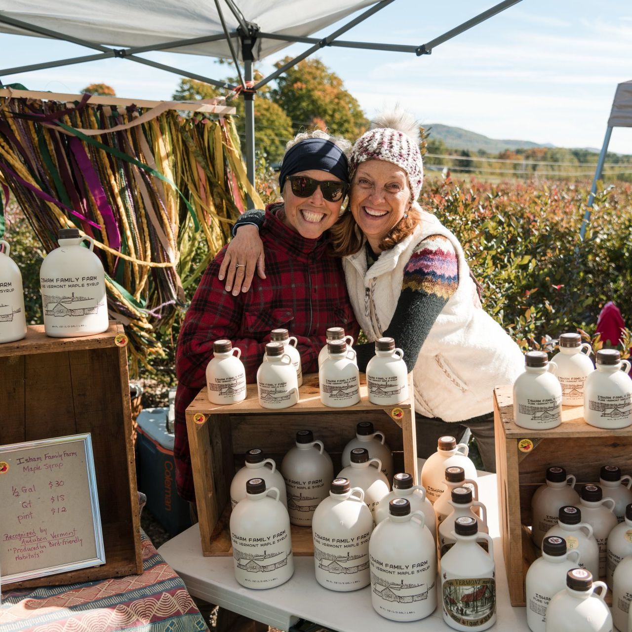 helen isham and friend behind the maple syrup table at isham family farmer's market in vermont