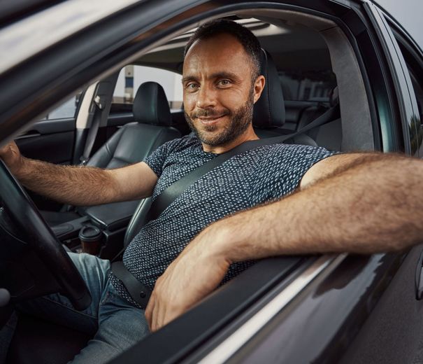 Man sitting in the driver’s seat of a car, smiling and resting one arm on the open window.