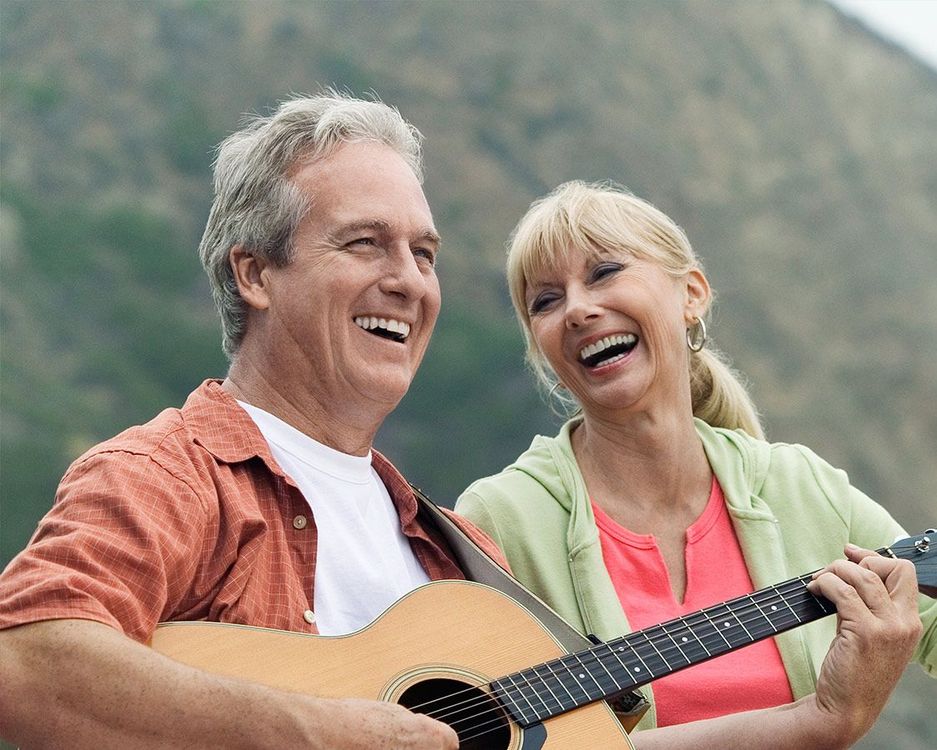 Smiling couple outdoors, laughing while playing guitar with mountains in the background