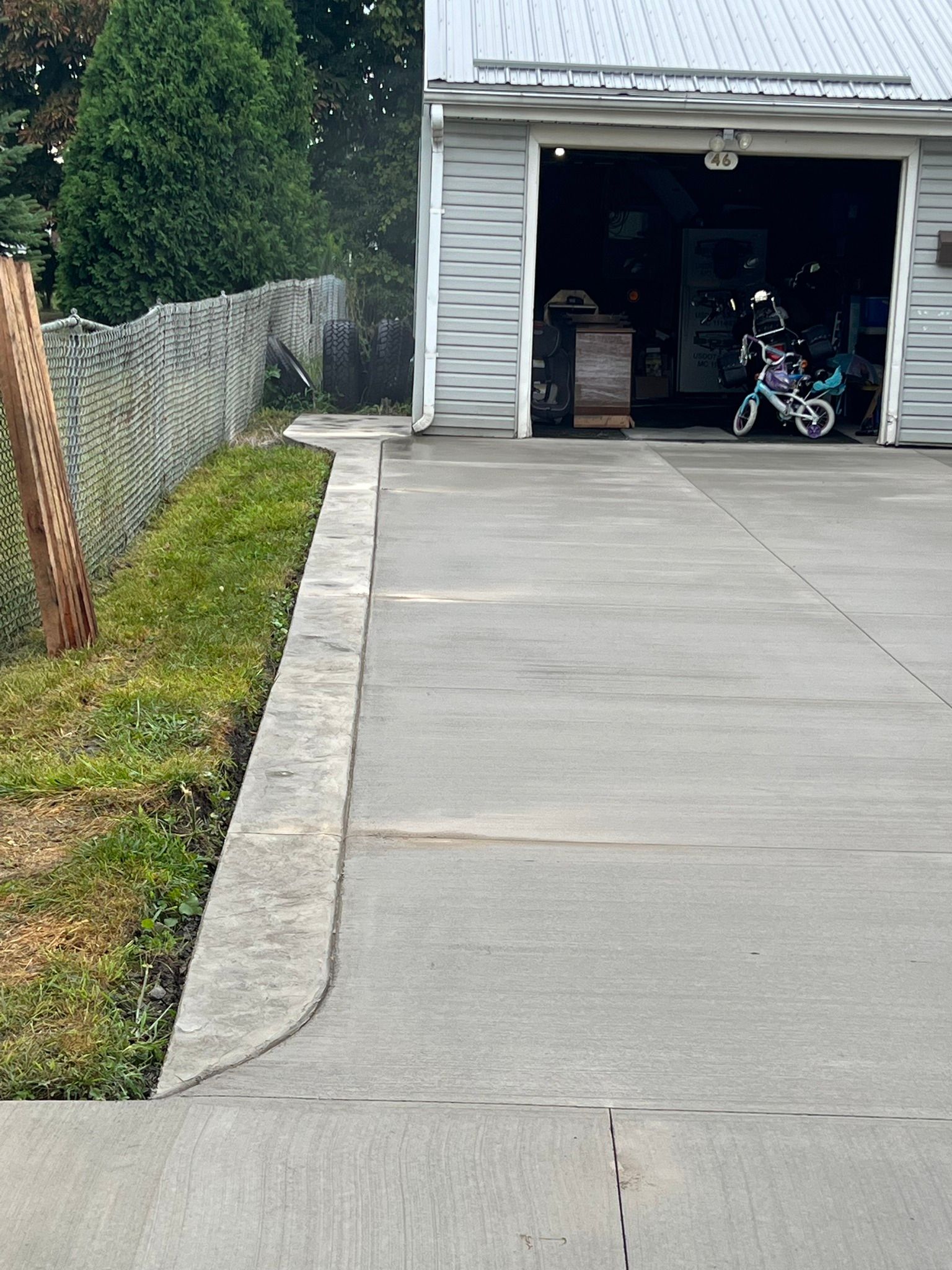 A concrete driveway leads to a garage with a person on a bicycle parked inside, alongside a grassy verge and a fence.