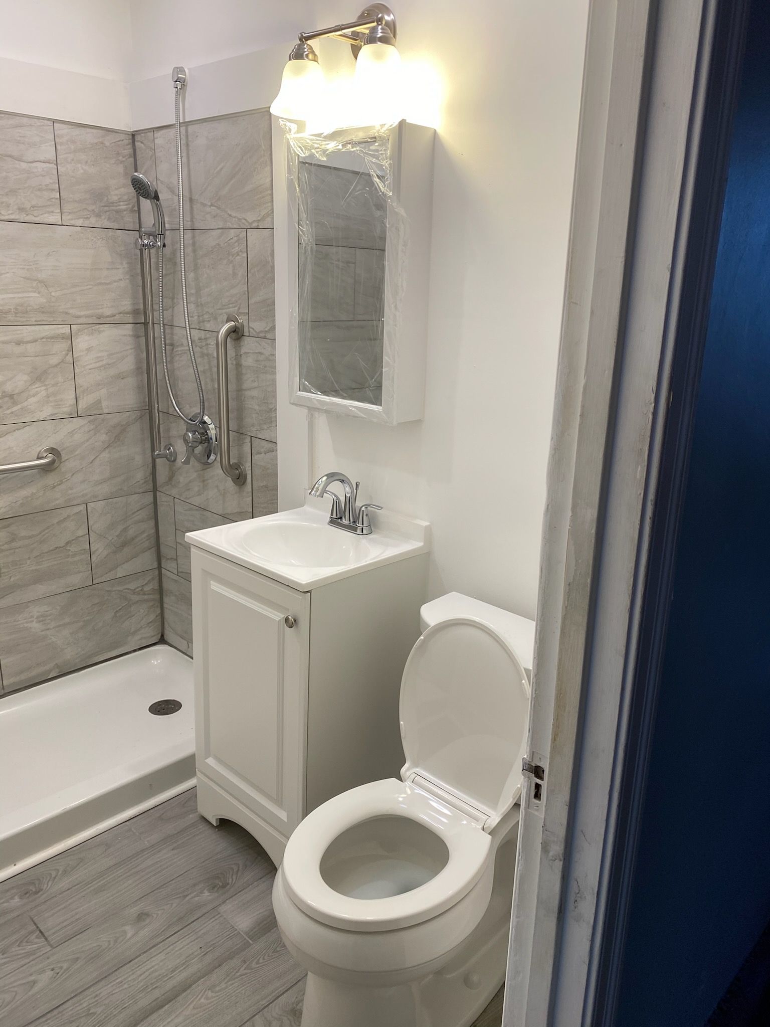 Bathroom with toilet, sink, shower with grab bar, and mirror. Gray and white color scheme.