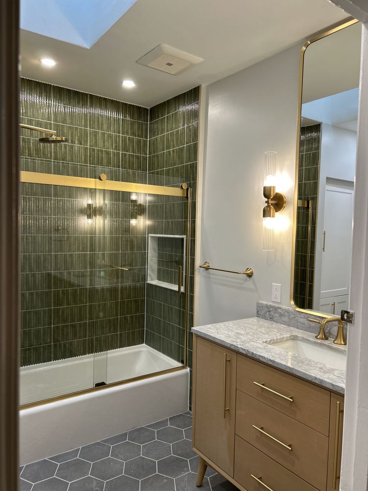 Bathroom with green tiled shower, gold fixtures, light wood vanity, and gray hexagon floor tiles.