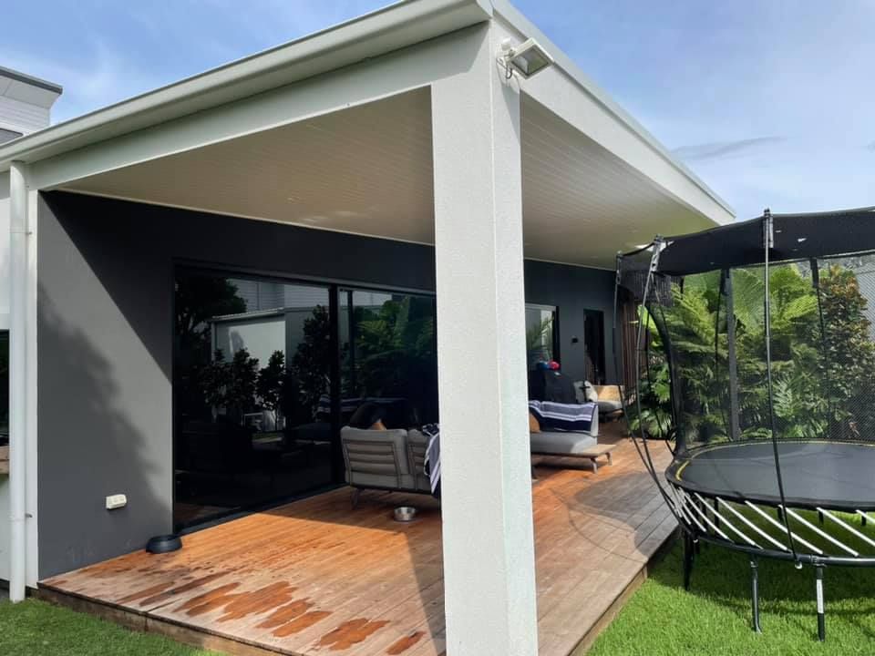 Outdoor covered patio with wooden deck, gray walls, and trampoline in the backyard.
