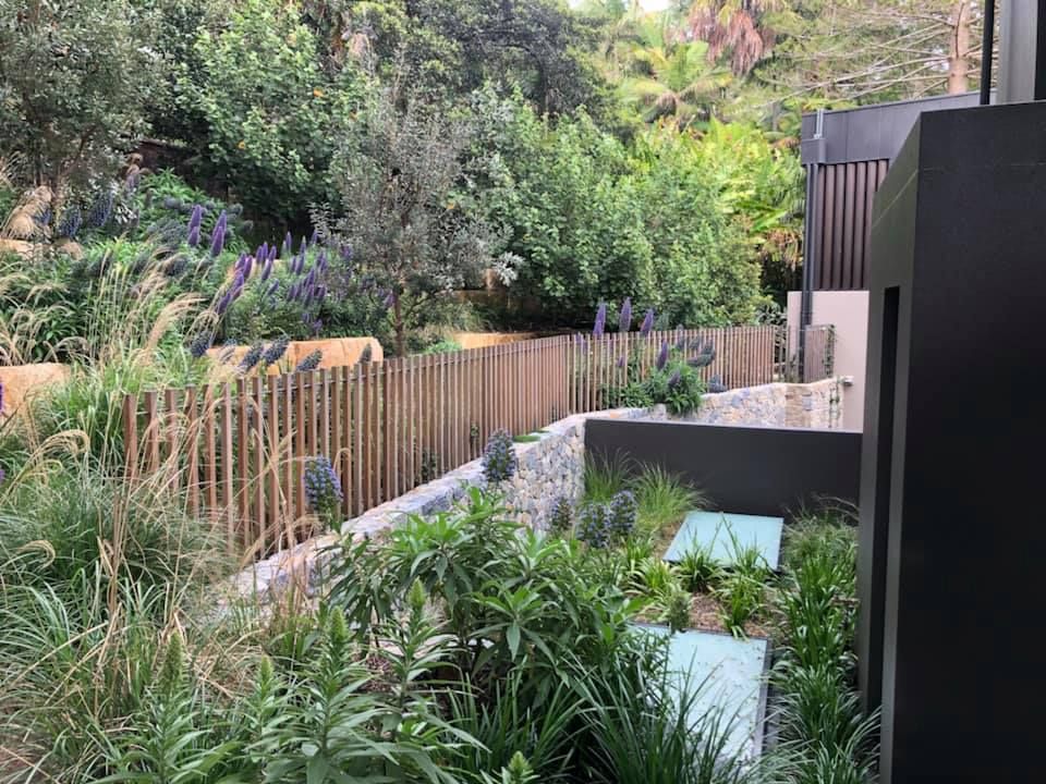 A tiered garden with wooden fence, stone walls, greenery, and a dark structure.