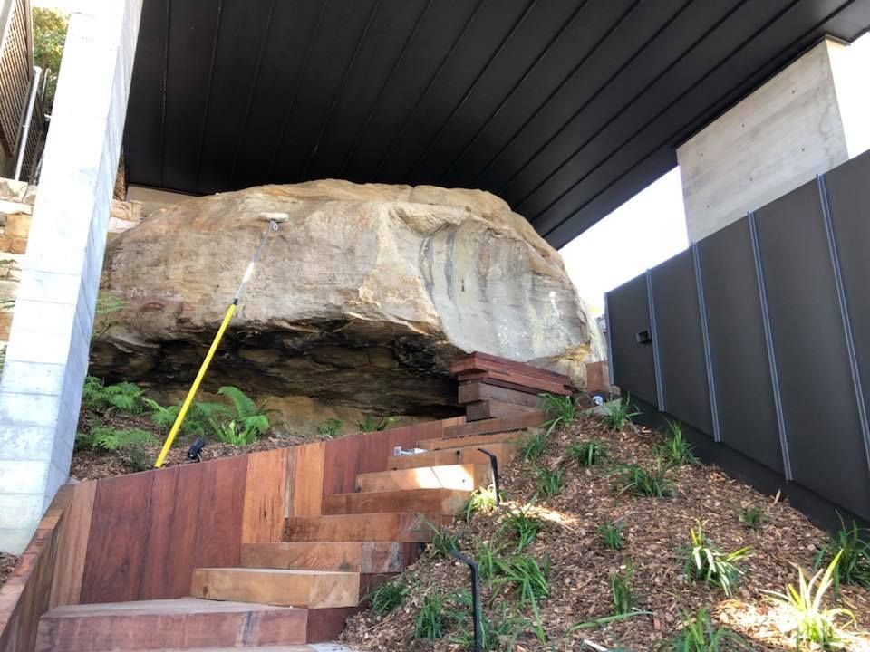 Wooden steps ascend a landscaped garden toward a large boulder under a dark overhang; metal fence on the right.