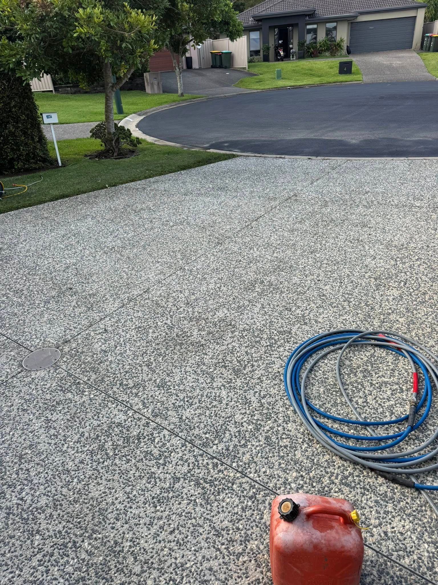 Gravel driveway with a coiled blue hose and red container in the foreground, house in the background.