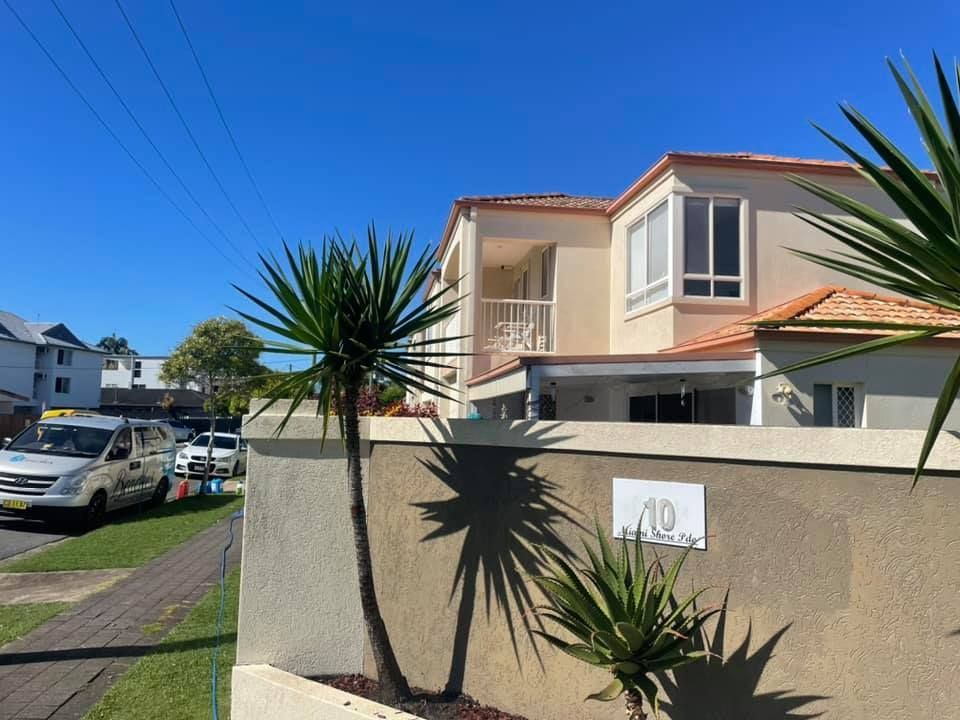 Cream-colored two-story building with a tiled roof, palm trees, and a low wall, under a bright blue sky.