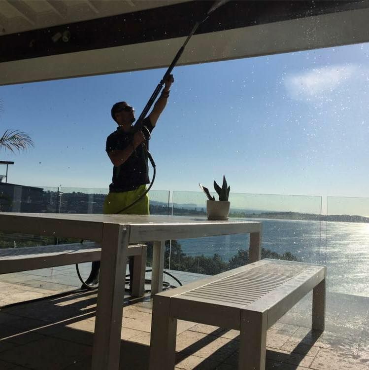 Person pressure washing a patio ceiling. Overlooks ocean on a sunny day. White table and bench in foreground.