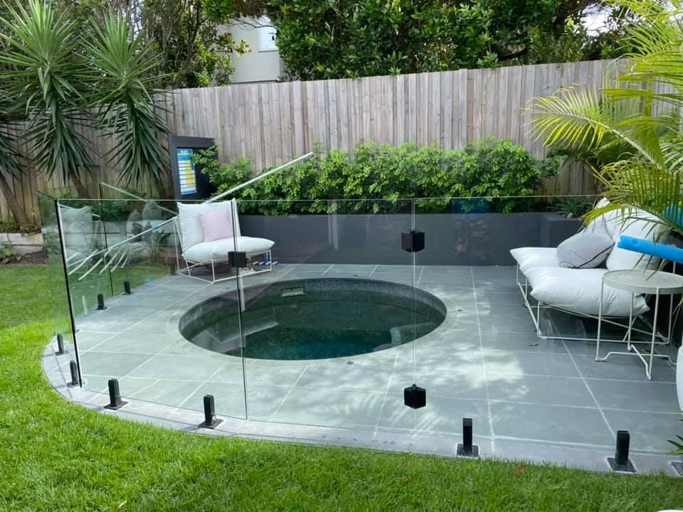 Backyard with a dark circular plunge pool, glass fence, lounge chairs, and lush greenery.