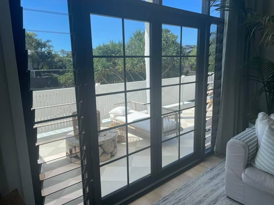 Black-framed glass doors open to an outdoor patio with white furniture, shutters, and a leafy background.