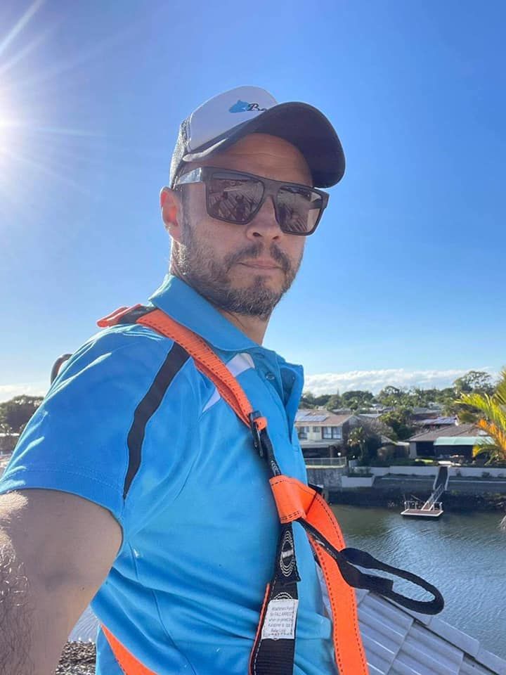 Man on rooftop wearing sunglasses and safety harness; sunny outdoor setting.