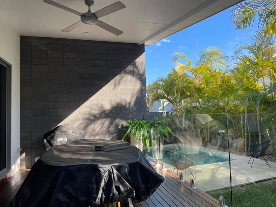 Covered outdoor table under a ceiling fan on a patio overlooking a pool and palm trees.