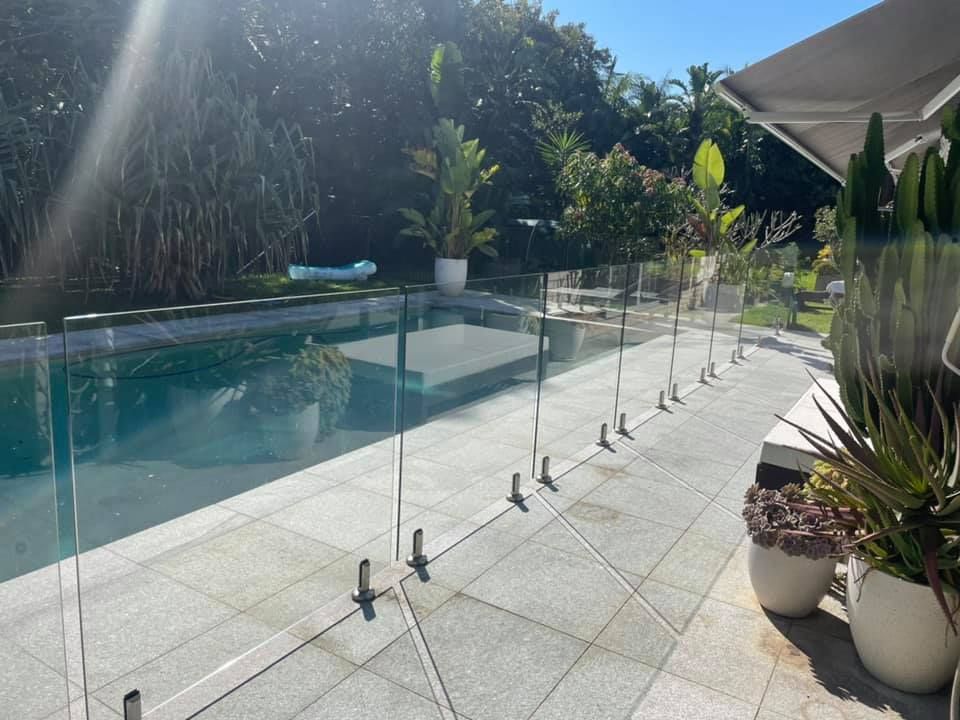 Glass fence surrounds a pool on a sunny patio with potted plants and lush foliage in the background.