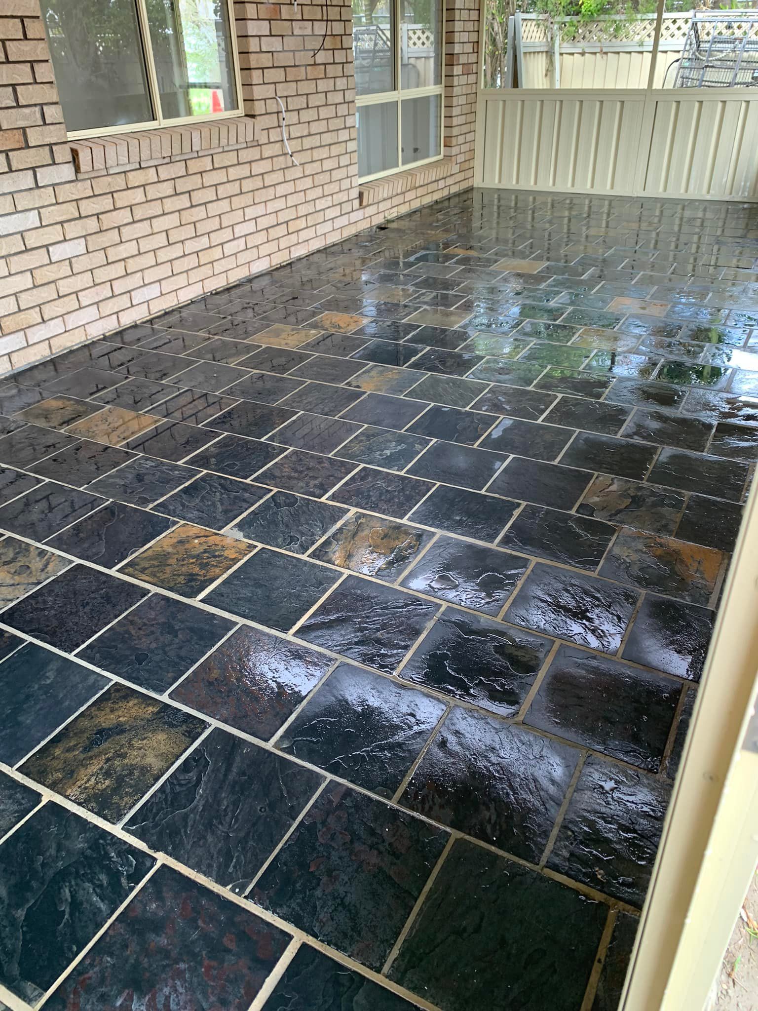 Wet, dark square-tiled patio with mixed tan tiles, under a brick wall and white fence.