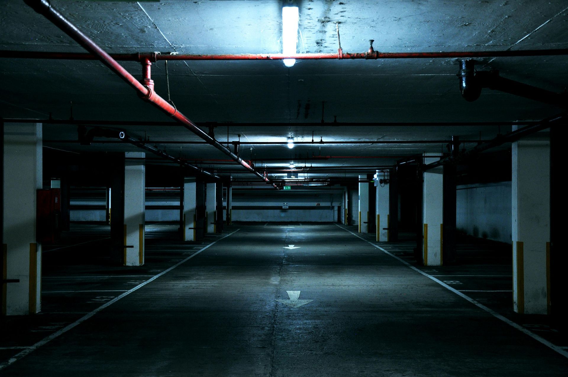 Empty, dimly lit underground parking garage. Concrete columns, red pipes, white lines on the floor, overhead lights.