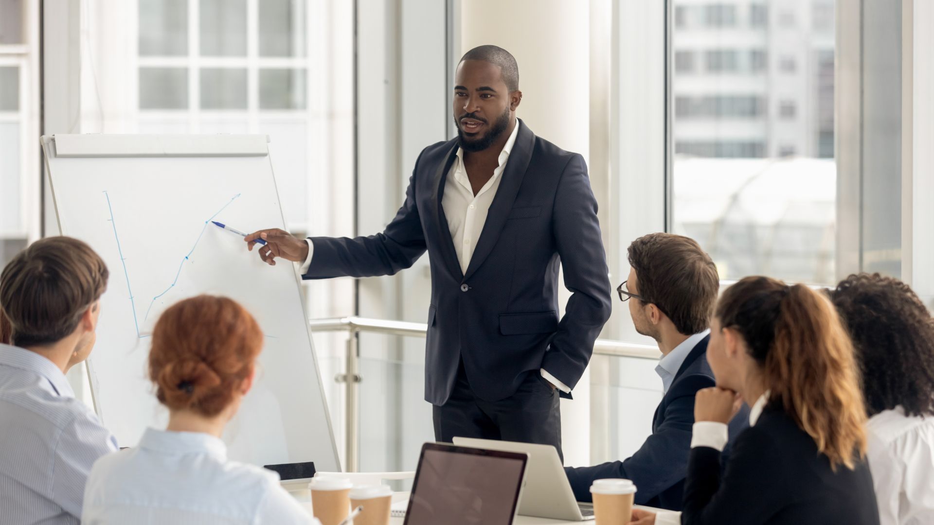 Man in suit presents a graph to a diverse group in a modern office, pointing with a pen.
