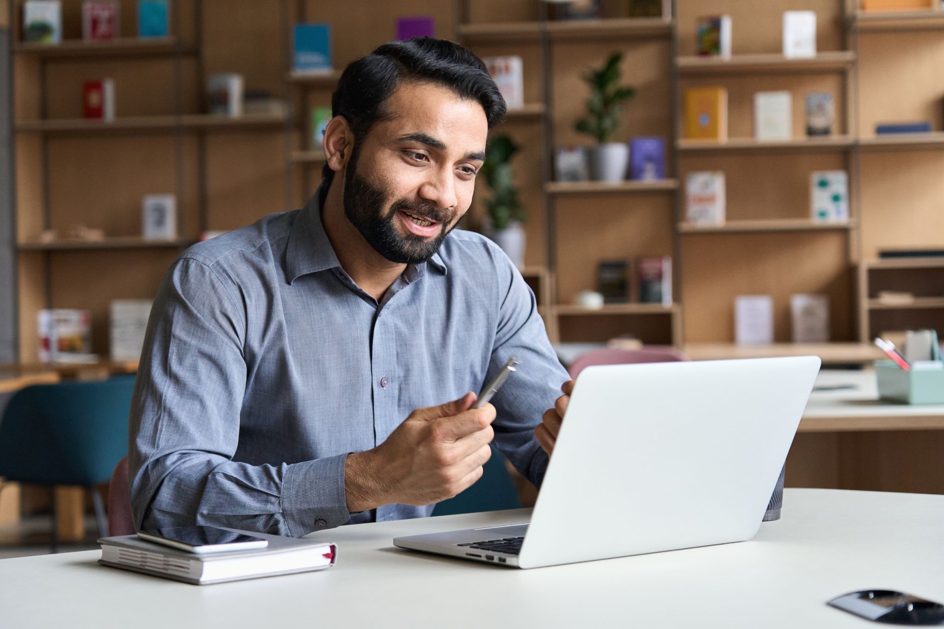 Man with beard smiles while on a video call using a laptop at a table, bookshelves in background.