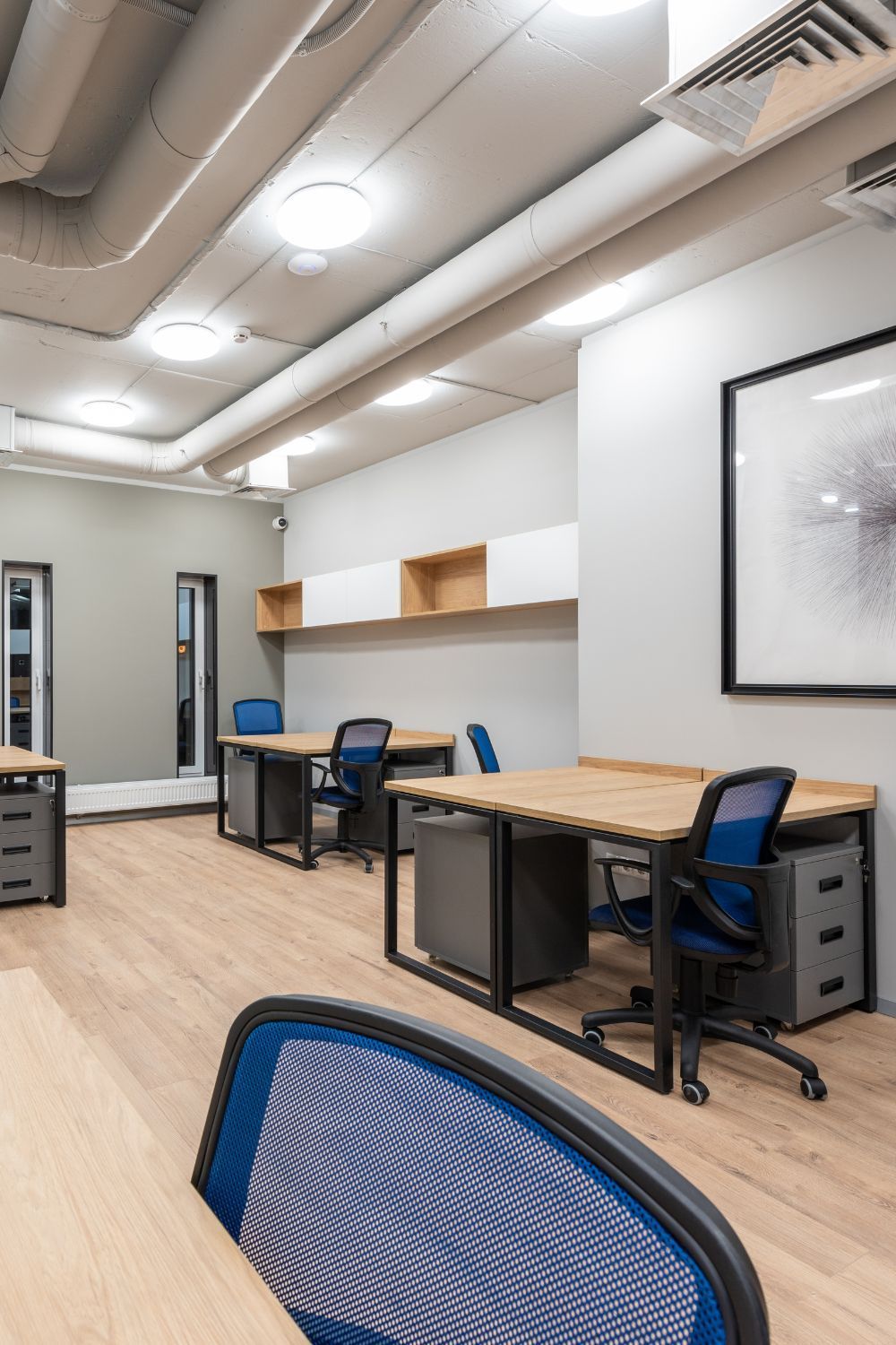 Office workspace with desks, blue chairs, and neutral walls. Lighting and artwork present.