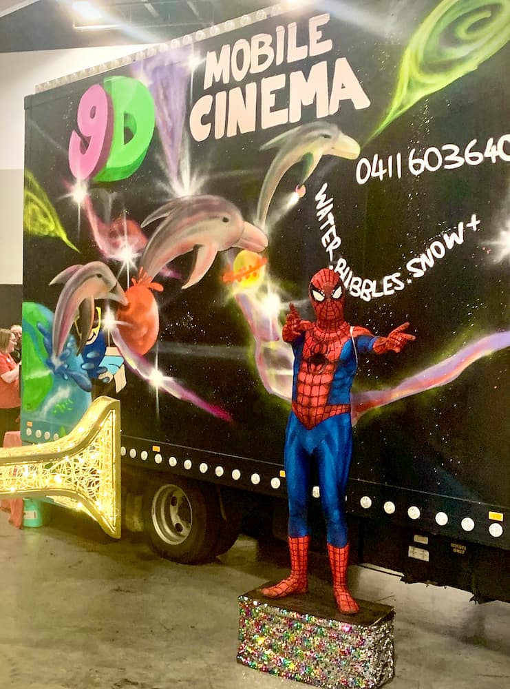 A statue of spider man is standing in front of a mobile cinema truck.