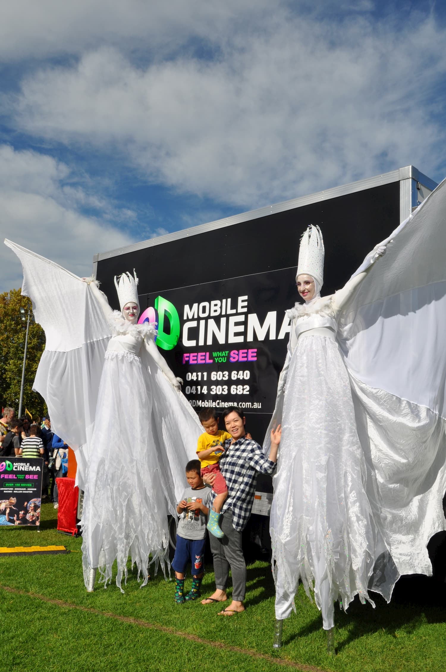 Two women in white stilts are standing in front of a mobile cinema truck.