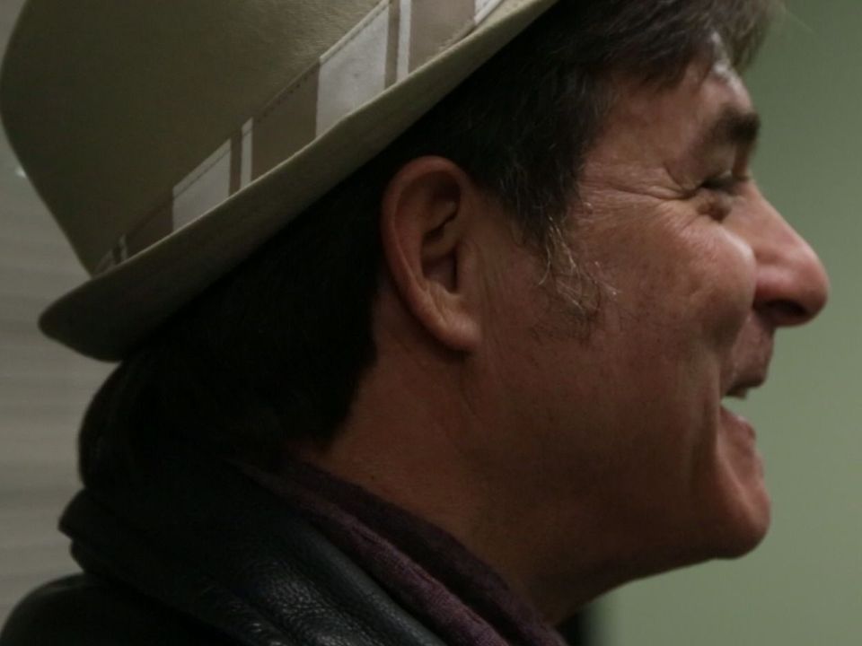 Man with a fedora hat, smiling in profile. Brown hair, leather jacket, neutral background.